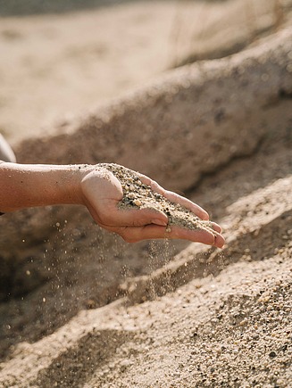 Nahaufnahme einer Hand, die natürlichen Sand mit feinen Kieseln auf einer Sandfläche rieseln lässt.