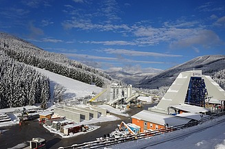Mobile concrete mixing plant with several silos, conveyor belts and aggregate boxes on a large construction site in a snow-covered alpine environment for concrete production.