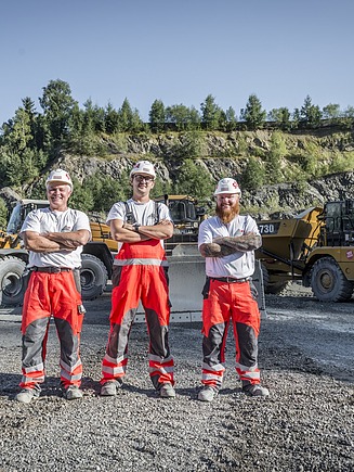 Three people in work clothes are standing on a gravel-covered area in front of a wheel loader and a dump truck in a quarry; layers of rock and vegetation are visible in the background.