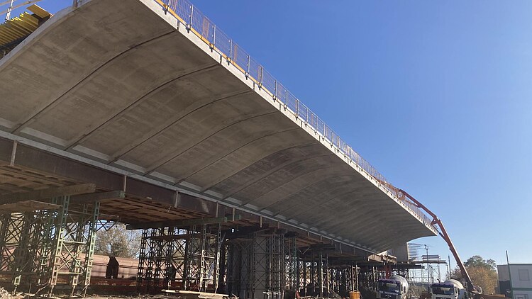 A concrete mixer truck is delivering concrete to a building site, whilst a concrete pump is feeding the fresh concrete via a boom onto a bridge under construction, which features formwork and a steel structure.
