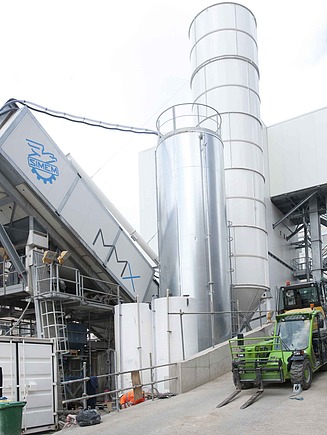 Mobile concrete mixing plant with inclined mixing module, high cement silo, technical containers and conveyor vehicle on a large inner-city construction site between multi-storey buildings.
