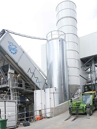Mobile concrete mixing plant with inclined mixing module, high cement silo, technical containers and conveyor vehicle on a large inner-city construction site between multi-storey buildings.