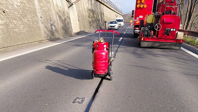 Risssanierung auf einer Asphaltfahrbahn, bei der ein längs verlaufender Straßenriss mit erhitztem bituminösem Dichtstoff maschinell verfüllt wird.