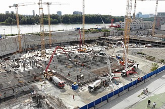 Großbaustelle mit mehreren Betonpumpen und Fahrmischern, die Frischbeton auf eine weitläufige, bewehrte Fundamentfläche einbringen, umgeben von Kränen und Baugruben in städtischer Lage.
