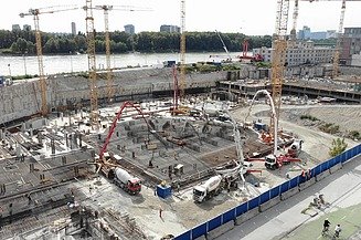 A large construction site with several concrete pumps and truck mixers pouring fresh concrete onto an extensive, reinforced foundation area, surrounded by cranes and excavation pits in an urban setting.
