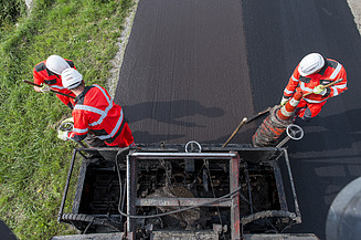 Two people in protective clothing work with hand tools on a freshly applied asphalt surface; a road construction machine is visible in the foreground