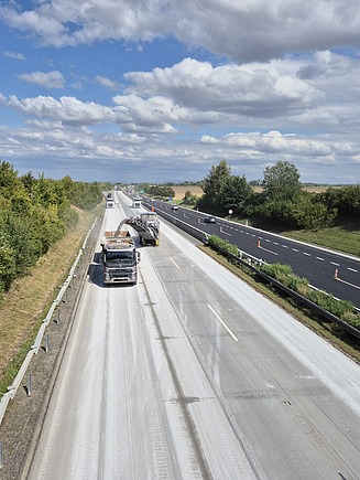 Autobahnbaustelle mit Fräsmaschine und Lkw auf einer Fahrbahn, während der Verkehr auf der benachbarten Spur weiterläuft; umgeben von Bäumen und offener Landschaft.