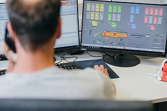 A person is working at a computer workstation with monitors displaying the control system and mix design for a concrete batching plant