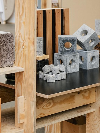 Ausstellung mit verschiedenen Baustoffproben wie Beton, Holz und Leichtmaterialien auf Regalen aus Holz, darunter Bauteile mit Öffnungen und strukturierte Materialblöcke.