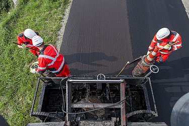 Two people in protective clothing work with hand tools on a freshly applied asphalt surface; a road construction machine is visible in the foreground