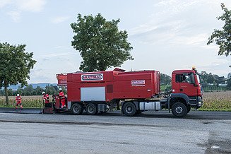 Multimac‑Spezialfahrzeug mit Bitumenemulsion im Einsatz auf einer Straße; mehrere Personen führen Instandhaltungsarbeiten durch