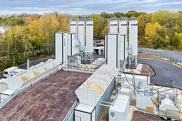 Mobile concrete mixing plant with four high cement silos, aggregate boxes, conveyor technology and technical containers on a paved area, surrounded by autumnal trees.