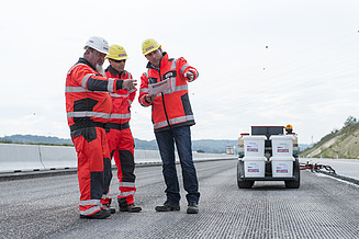 Technical advice on a road construction site, where specialised personnel in protective clothing discuss construction plans and coordinate the use of bituminous products on site.