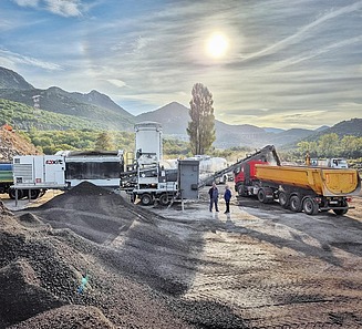 Anlage für Kaltrecycling in plant verarbeitet aufbereitetes Asphaltmaterial auf einer Baustelle; Förderband verlädt das Recyclinggut in Lkw, während Tankfahrzeuge und Maschinen im Hintergrund stehen