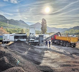 A cold recycling plant processes pre-treated asphalt material at a construction site; a conveyor belt loads the recycled material onto lorries, whilst tankers and machinery are parked in the background