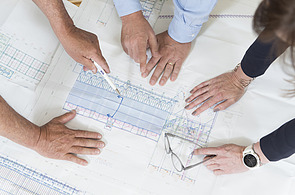 Several hands work together on large-format technical drawings for the design and plant planning of mobile concrete mixing plants; the focus is on detailed construction plans for concrete and plant components on large construction sites.