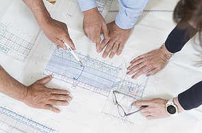 Several hands work together on large-format technical drawings for the design and plant planning of mobile concrete mixing plants; the focus is on detailed construction plans for concrete and plant components on large construction sites.