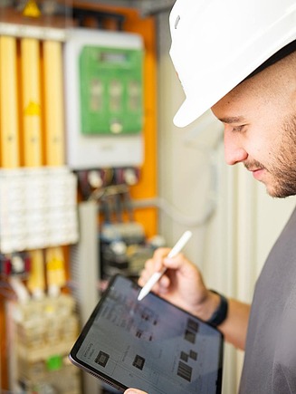 Person mit Schutzhelm bedient ein Tablet vor einem geöffneten Schaltschrank mit elektrischen Komponenten, Leitungen und technischen Modulen.