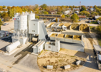 Luftaufnahme einer Betonmischanlage mit mehreren Silos, Zuschlagstofflagern, Förderanlagen und Fahrzeugen auf einem Betriebsgelände.