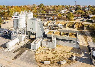 Aerial view of a concrete mixing plant with several silos, aggregate storage areas, conveyor systems and vehicles on the site.