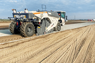 Straßenrecyclingmaschine verarbeitet vorhandenes Material auf einer Fahrbahn und verteilt es gleichmäßig, während eine zweite Maschine im Hintergrund die Oberfläche weiterbearbeitet.