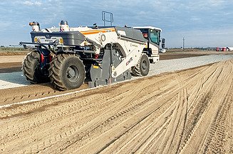 Straßenrecyclingmaschine verarbeitet vorhandenes Material auf einer Fahrbahn und verteilt es gleichmäßig, während eine zweite Maschine im Hintergrund die Oberfläche weiterbearbeitet.