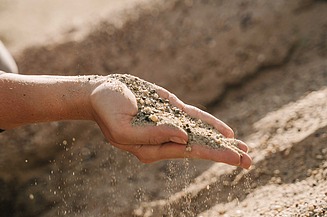 Hand hält Sand und feine Gesteinskörnung, die zwischen den Fingern rieselt, im Hintergrund ist weiteres Schüttmaterial sichtbar.