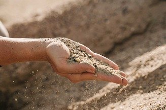 Hand hält Sand und feine Gesteinskörnung, die zwischen den Fingern rieselt, im Hintergrund ist weiteres Schüttmaterial sichtbar.