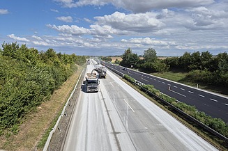 Autobahnbaustelle mit Fräsmaschine und Lkw auf einer Betonfahrbahn, während der Verkehr auf der benachbarten Spur weiterläuft