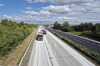 Autobahnbaustelle mit Fräsmaschine und Lkw auf einer Betonfahrbahn, während der Verkehr auf der benachbarten Spur weiterläuft