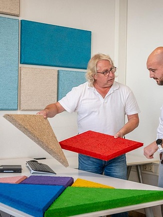 Two people look at and compare different coloured fibre-based building boards on a table in an interior room with attached wall panels.