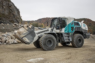 A wheel loader bearing an H₂ logo is moving rock in a quarry; rock faces and stockpiled material can be seen in the background.