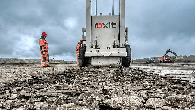 Straßenbaumaschine verarbeitet aufgebrochenes Asphalt- und Gesteinsmaterial auf einer Baustelle, während Personen in Schutzkleidung daneben stehen