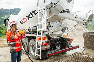 A concrete mixer is unloading concrete via a discharge chute at a construction site, whilst the machine is being cleaned with a water hose