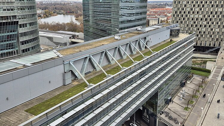 Oblique view of a large office and technical building with an elongated roof surface on which several green sections are arranged between technical superstructures and railings, surrounded by high-rise buildings in an urban environment.