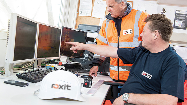 Two people are looking at several monitors displaying technical plant and process visualisations; one person is pointing at the screen, with input devices and a safety helmet in the foreground